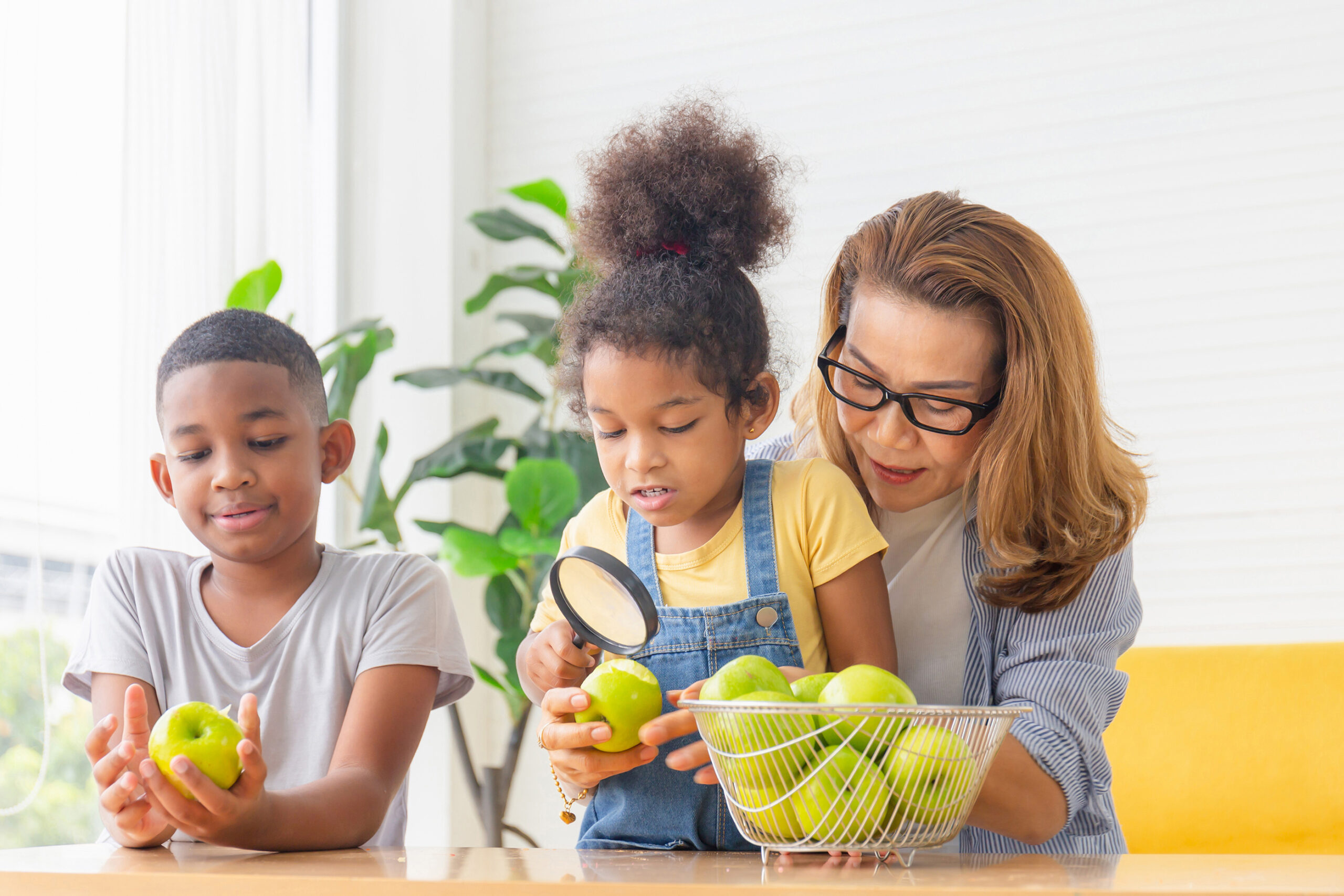 A woman and her grandchildren smiling and looking at fruit with a magnifying glass